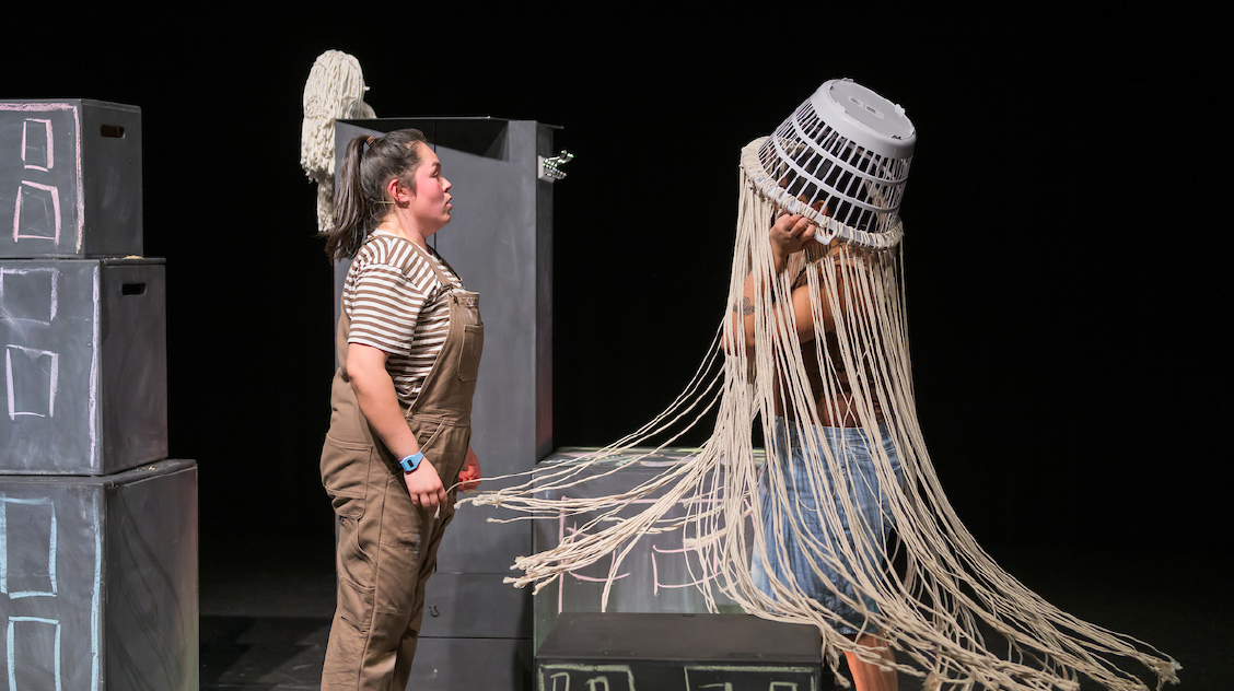 Performers on stage, one wearing brown overalls and the other wearing a laundry basket prop on her head with long rope for hair.