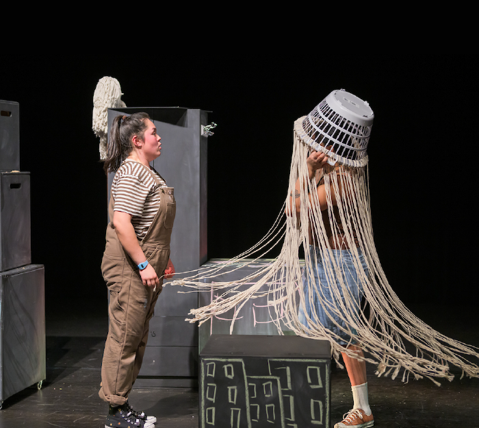 Performers on stage, one wearing brown overalls and the other wearing a laundry basket prop on her head with long rope for hair.