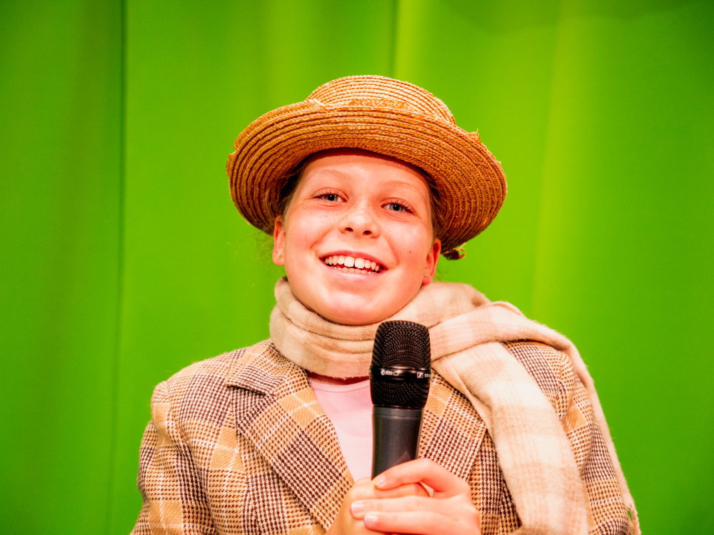 A child standing in front of a greenscreen, holding a microphone.